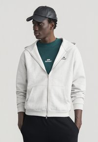 Νεαρός άνδρας φοράει μαύρο καπέλο, μπλουζάκι με λογότυπο σε τιρκουάζ χρώμα, ανοιχτό γκρι φούτερ με φερμουάρ και μαύρο παντελόνι, στέκεται με τα χέρια στις τσέπες κοιτάζοντας πλάγια.