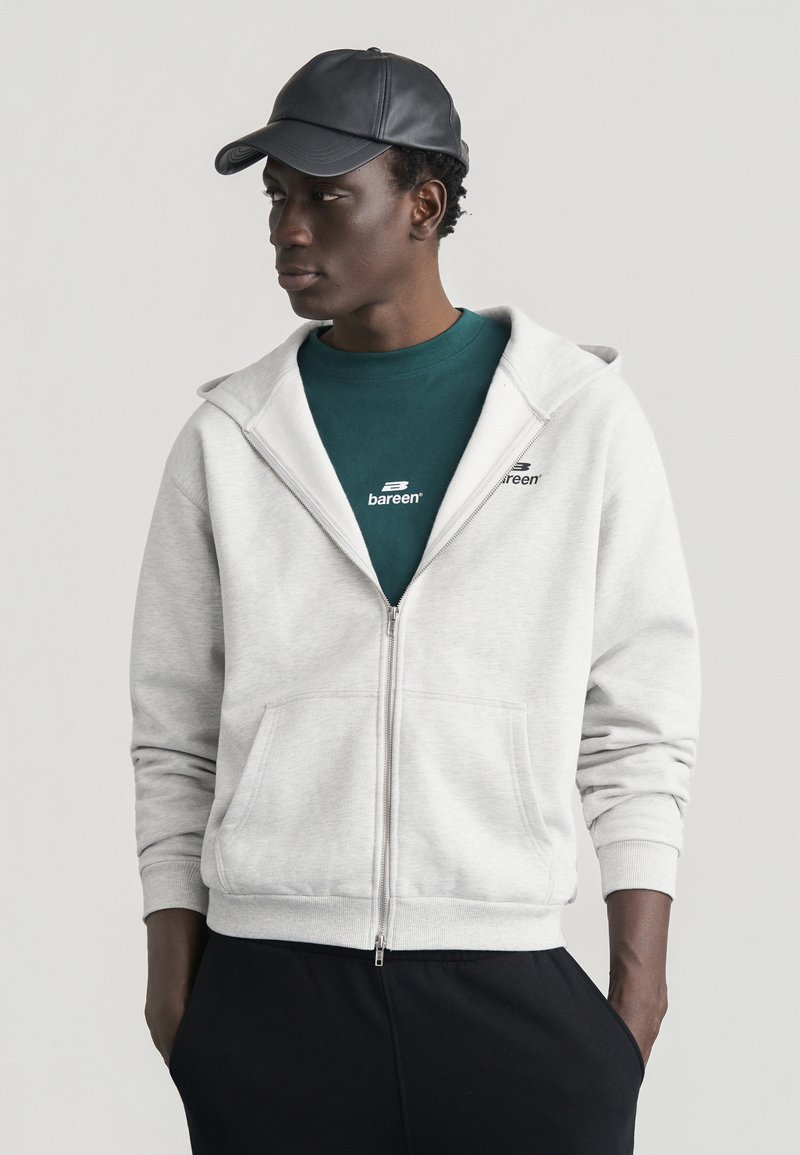 Νεαρός άνδρας φοράει μαύρο καπέλο, μπλουζάκι με λογότυπο σε τιρκουάζ χρώμα, ανοιχτό γκρι φούτερ με φερμουάρ και μαύρο παντελόνι, στέκεται με τα χέρια στις τσέπες κοιτάζοντας πλάγια.