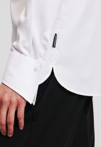 Witte longsleeve shirt met een knoopmanchet en een logo label aan de zijkant. Glad materiaal met een getailleerde snit en een afgeronde zoom.