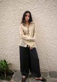 Hellbeige strukturierte Bluse mit Kragen und langen Ärmeln, kombiniert mit locker sitzenden schwarzen Hosen und flachen schwarzen Schuhen, vor einer Steinewand.