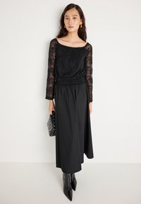 Μαύρη μπλούζα με μακριά μανίκια και φλοράλ δαντέλα, συνδυασμένη με μια μαύρη, ρο(flow) μάξι φούστα. Το μοντέλο κρατάει μια μαύρη τσάντα με τρουκς. Μαύρες μυτερές μπότες.