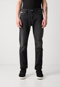 Schwarze Jeans aus Denim mit geradem Schnitt, verblasster Optik, Fünf-Taschen-Design und Metallbeschlägen am Bund. Getragen mit schwarzen Sneakers.