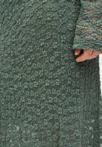 Rankos uždėjimo ant žalios gėlių nėrinių medžiagos, kurioje iškilę gėlių raštai ir tinklinis fonas, artimas planas.