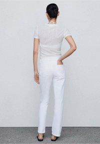 Top blanco transparente de manga corta con un detalle de lazo en la parte posterior, combinado con jeans rectos blancos con bolsillos traseros y costuras mínimas.