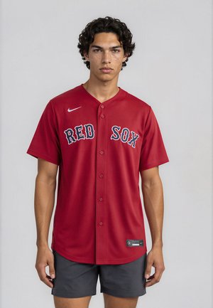 Jonge man met krullend haar die een rood Red Sox honkbalshirt en zwarte korte broek draagt, staande tegen een effen lichte achtergrond.