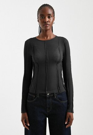Vrouw met gevlochten haar, gekleed in een getailleerde zwarte geribbelde lange mouwen top en donkerblauwe spijkerbroek, staand tegen een effen lichte achtergrond.