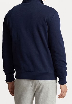 Homme portant un sweat-shirt à manches longues bleu marine et un pantalon de survêtement gris clair, vu de dos sur un fond uni.