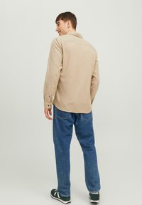 Beige Langarmhemd mit knöpfbarem Kragen, strukturiertem Stoff, geradem Schnitt; kombiniert mit blauen Jeans und dunklen Sneakers mit weißen Akzenten.