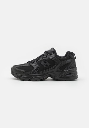 Baskets de sport noires avec lacets à l'avant, col rembourré et logo "N" estampillé sur le côté, présentées de profil sur fond blanc.