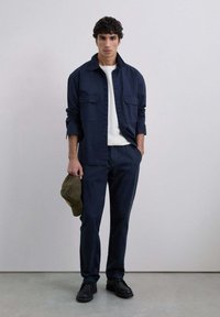 Chaqueta de camisa azul marino con dos bolsillos frontales, combinada con pantalones azul marino a juego y zapatos negros. Sosteniendo una gorra de cuadros verde.