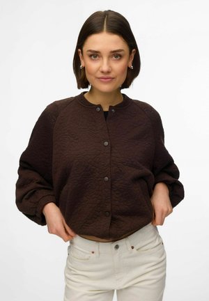 Jeune femme portant une veste matelassée marron foncé à boutons et un jean de couleur claire, ajustant l'ourlet de sa veste sur un fond blanc uni.