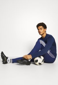 Marineblauwe sportieve longsleeve top en broek met drie witte strepen. Zwarte voetbalschoenen met het label "NEMESIS" en een zwart-witte voetbals. 