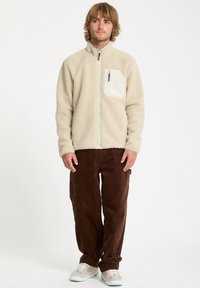 Veste en polaire couleur crème avec un col montant et une poche avant, associée à un pantalon en velours côtelé marron et des baskets claires.