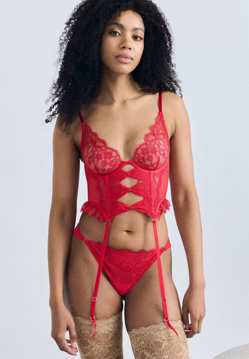 Σετ κόκκινης δαντελένιας εσώρουχα με κορσέ που έχει λουλουδένια σχέδια, ρυθμιζόμενες τιράντες και βολάν, σετ με τόνγκα που ταιριάζει και διαφανείς κάλτσες.