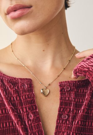 Gold heart-shaped pendant necklace with a textured chain, worn over a deep red, ribbed fabric top featuring a button detail.