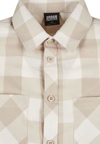 Chemise à carreaux beige et blanche avec boutons, col, poches poitrine, et étiquette Urban Classics indiquant la taille XS, fabriquée en Chine.