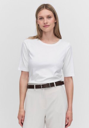 Vrouw met steil blond haar, gekleed in een wit shirt met korte mouwen, beige broek en een donkerbruine riem, staand tegen een effen achtergrond.