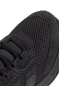 Gros plan d’une chaussure de sport noire avec tissu en mesh, superpositions texturées et laçage à l’avant sur fond blanc.