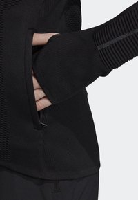 Haut de sport noir en tissu tricoté texturé, avec poche latérale et trous pour les pouces dans les manches pour couvrir les mains. Conçu pour le confort et la liberté de mouvement.