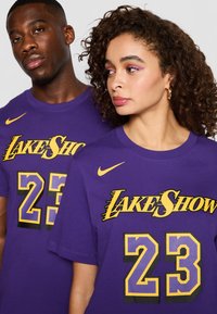 Fialová bavlněná trička mají žlutý nápis "LAKE SHOW" a číslo "23". Logo Nike je umístěno na levém rameni. Unisex design.