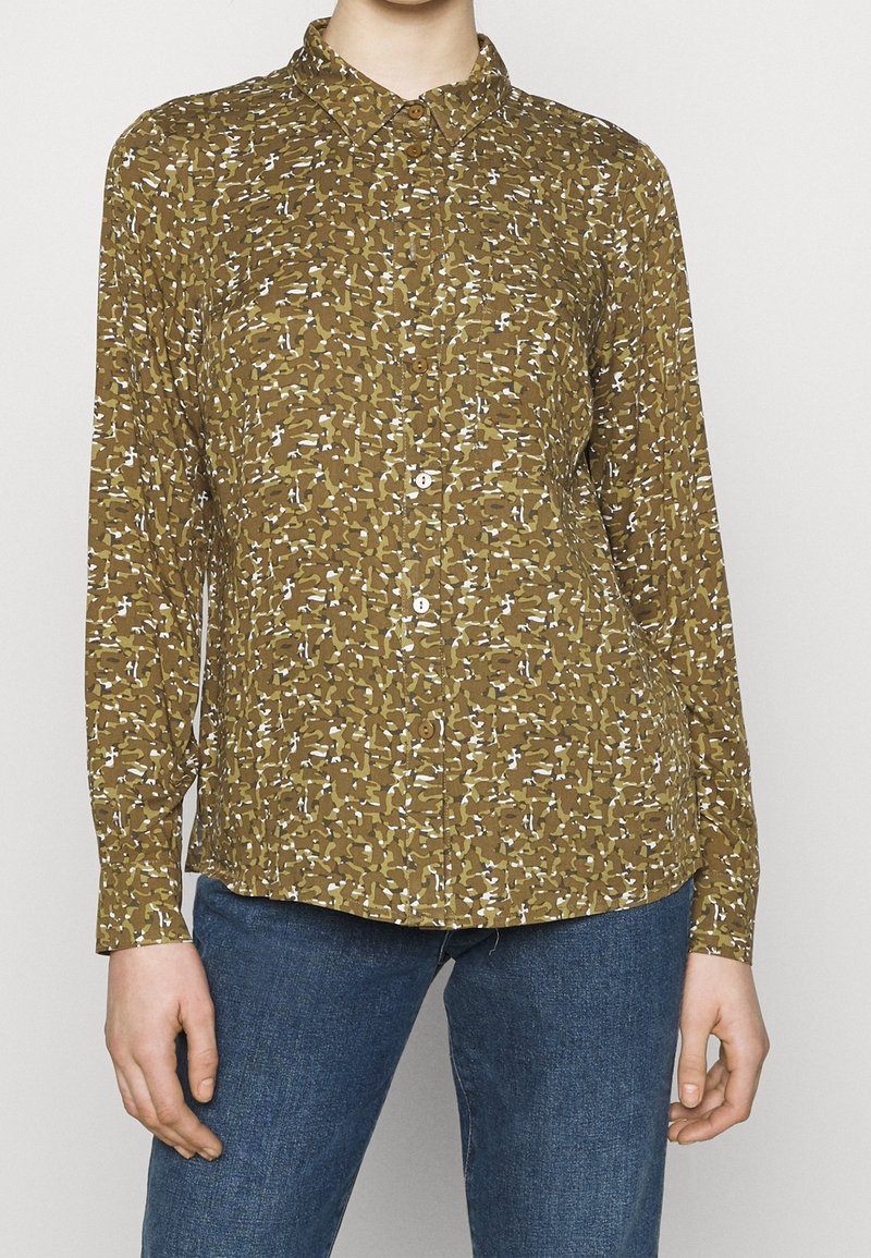 Femme porte une chemise boutonnée à manches longues avec un motif abstrait marron, blanc et noir, associée à un jean en denim bleu.
