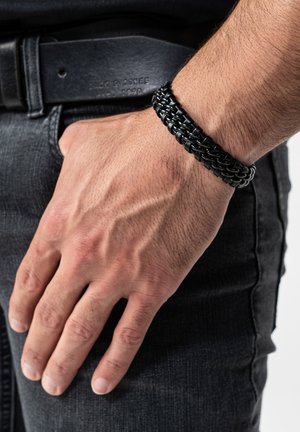 Black woven bracelet made of rubber, featuring a thick, braided pattern with a smooth texture. Worn on a wrist, paired with dark clothing.