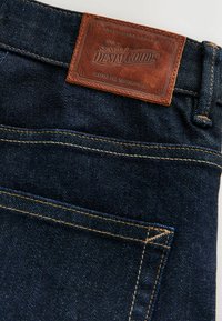 Tumši zili denim džinsi ar redzamu šuvju un brūnu ādas birku, kas ar šuvēm piestiprināta pie jostasvietas, uz kuras rakstīts "Signature Denim Goods".