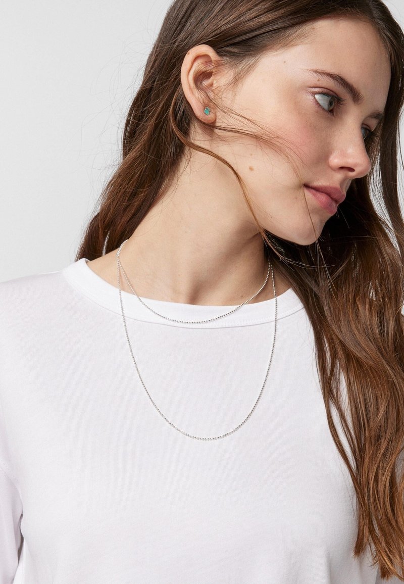 Collar de cadena de plata con pequeñas cuentas redondas que caen alrededor del escote; modelo vistiendo una camisa blanca sencilla y con el cabello largo y castaño.