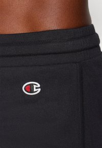 Närbild av en persons midja iförd svarta sweatpants med en broderad röd, vit och svart Champion-logga på höftområdet.