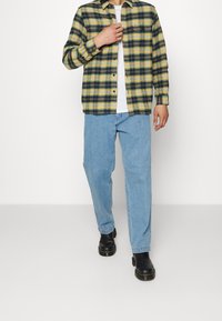 Rutig flanellskjorta i gult, grönt och svart, knäppt, med två bröstfickor och en logotags. Kombinerad med ljusblå jeans och svarta stövlar.