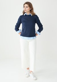 Granatowy sweter z długim rękawem nałożony na jasnoniebieską koszulę w paski, zestawiony z białymi spodniami o szerokich nogawkach i białymi tenisówkami.