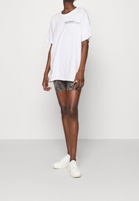 T-shirt bianca oversize con grafica testuale, abbinata a pantaloni corti grigi a fantasia. Modello indossa sneakers bianche, in piedi su uno sfondo neutro.