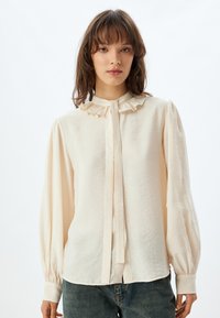 Blusa crema con colletto arricciato, maniche lunghe a palloncino, polsini con bottone e chiusura a bottoni; realizzata in tessuto leggero e strutturato.