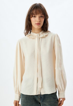 Blusa crema con colletto arricciato, maniche lunghe a palloncino, polsini con bottone e chiusura a bottoni; realizzata in tessuto leggero e strutturato.