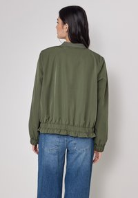 Giacca leggera verde oliva con polsini e orlo elasticizzati, caratterizzata da un design semplice. Abbinata a jeans in denim blu a gamba larga.