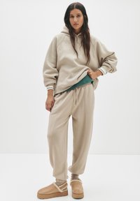 Beige übergroßer Hoodie mit Rippbündchen, kombiniert mit lockeren beigen Jogginghosen. Die Schuhe bestehen aus tanfarbenen Plattform-Clogs mit flauschigen Akzenten.