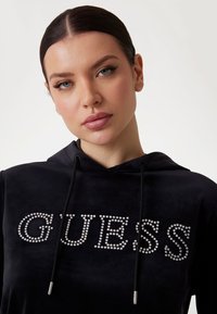 Čierna zamatová mikina s kapucňou, ktorá obsahuje kamienkový nápis "GUESS", s nastaviteľnou kapucňou a hladkou textúrou. Prvky zahŕňajú strieborné kovanie na šnúrkach.
