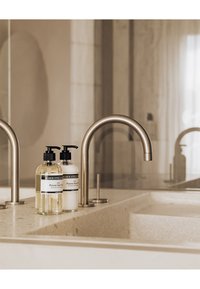 Due bottiglie di vetro trasparente con pompe nere, piene di un liquido e di una crema, su un bancone in pietra chiara accanto a un rubinetto in metallo curvo.