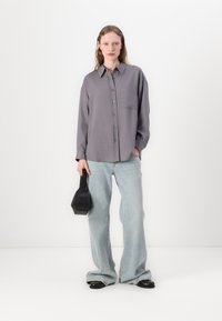 Camisa cinza de botones com um bolso no peito, combinada com jeans flare azul claro. Modelo segura uma bolsa geométrica preta, de pé contra um fundo neutro.