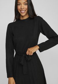Black knitted dress with long sleeves, round neckline, and a tied belt at the waist. Vertical ribbed texture throughout the fabric.