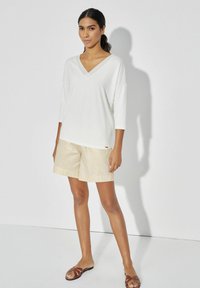 Lekki, długi rękaw, biały top z głębokim dekoltem w serek, w połączeniu z beżowymi, teksturowanymi szortami i brązowymi klapkami. Minimalistyczny design.