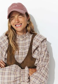 Casquette en velours côtelé rose poussiéreux avec le texte "LOVER", blouse à carreaux crémeuse froncée, et gilet en suédine marron. Manches bouffantes et détails de boutons visibles.