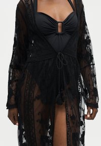 Maillot de bain noir froncé avec une découpe centrale, porté sous un voile en dentelle transparent à manches longues, noué à la taille avec des pompons.