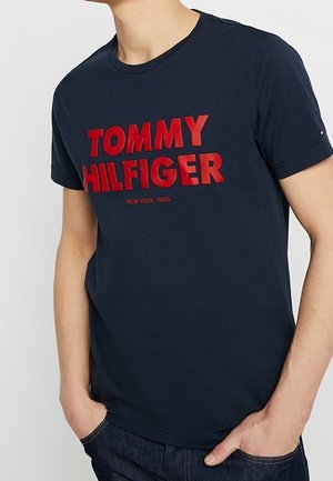 Personne portant un t-shirt bleu marine avec un texte rouge en gras "TOMMY HILFIGER" et un texte plus petit "NEW YORK, 1985" sur la poitrine.