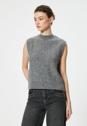 Grauer ärmelloser Strickpullover mit hohem Kragen, ausgestattet mit einem strukturierten Kabelmuster und einem gerippten Saum, kombiniert mit dunklen Jeans.