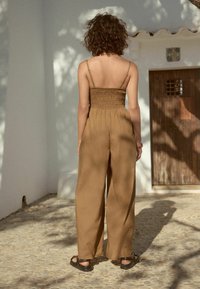 Brun, bredbygd jumpsuit med smockad bodice och tunna axelband. Sedd bakifrån, accessoriserad med svarta sandaler, mot en vit vägg.