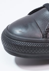 Gros plan sur la pointe d'une basket noire avec une semelle en caoutchouc texturée et un détail de coque en cuir lisse sur fond blanc.