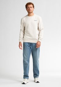 Felpa leggera beige con texture a puntini, collo rotondo e polsini a costine, abbinata a jeans blu a gamba dritta e sneakers bianche.