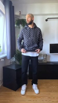 Muž s krátkymi blond vlasmi, oblečený v čiernej mikine so vzorom, čiernych nohaviciach a bielych teniskách, stojí v miestnosti s rastlinami a šijacím strojom.