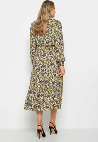 Vestido maxi de tela negra con estampado floral en amarillo, blanco y rosa. Mangas largas, cintura ceñida, falda plisada, combinado con tacones negros.